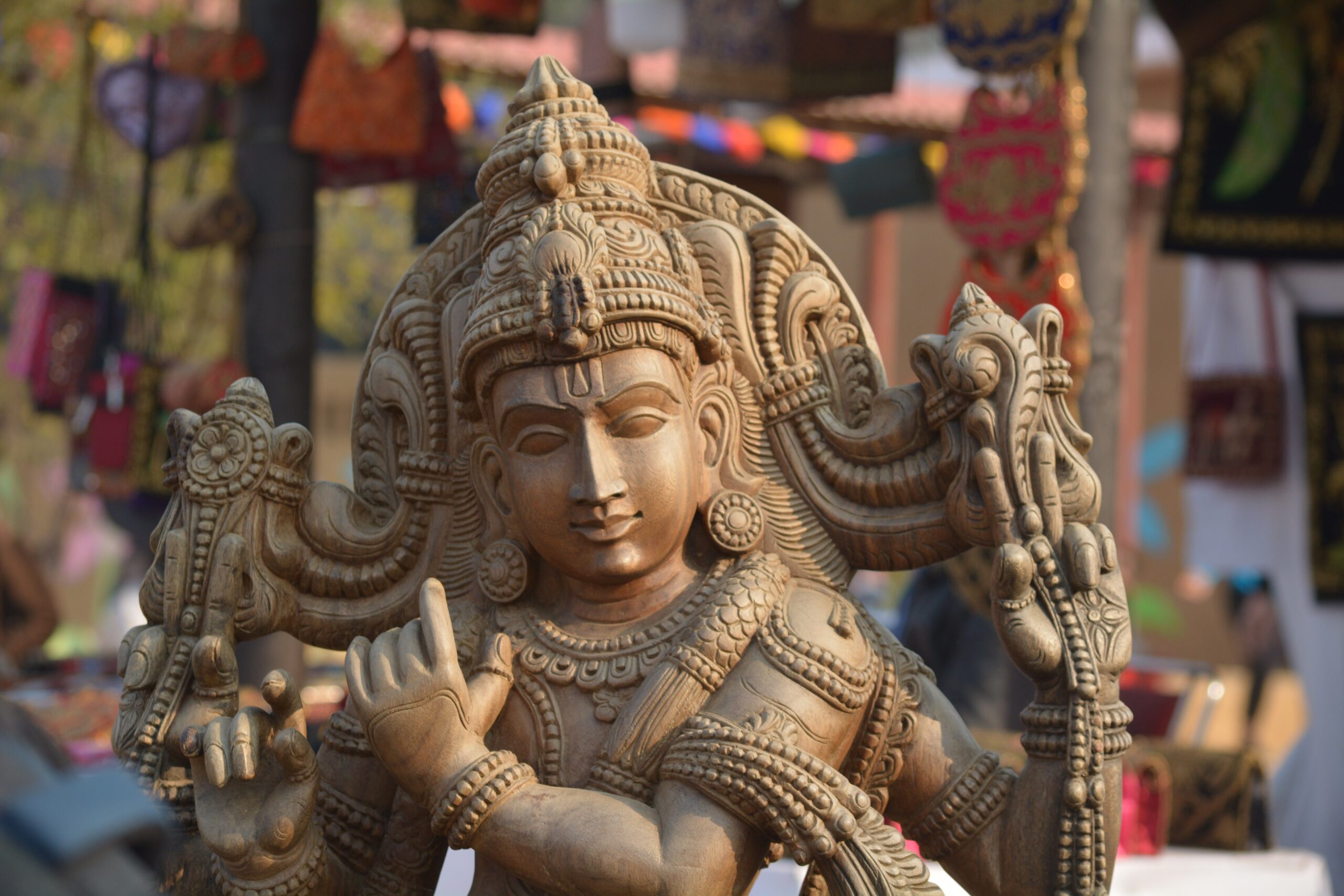 Intricately carved wooden statue of Lord Krishna with an ornate crown, multiple arms, and detailed jewelry, displayed at a craft market