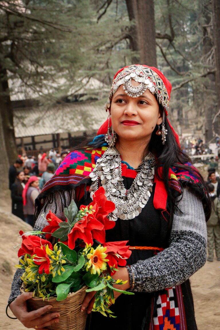 Woman dressed in traditional Himachali attire with silver coin jewelry, red headscarf, and embroidered shawl, holding a basket of flowers in a forest setting Caption: A woman dressed in traditional Himachali festive attire, adorned with silver jewelry and a vibrant headscarf Description: This image captures the rich cultural heritage of Himachal Pradesh through traditional attire worn during local festivals and celebrations. The look features an ornate silver headpiece and coin necklace, a bright red headscarf, and a colorful hand-woven shawl with geometric patterns, paired with a black dress. Holding a basket of vibrant red and yellow flowers, she stands amid deodar trees, reflecting the warmth and tradition of hill-state culture often seen during local fairs and festivals around Shimla. Let me know if you'd like this tied to a specific festival (like Shimla's Summer Festival) or location, and I can adjust accordingly. Woman dressed in traditional Himachali attire with silver coin jewelry, red headscarf, and embroidered shawl, holding a basket of flowers in a forest setting