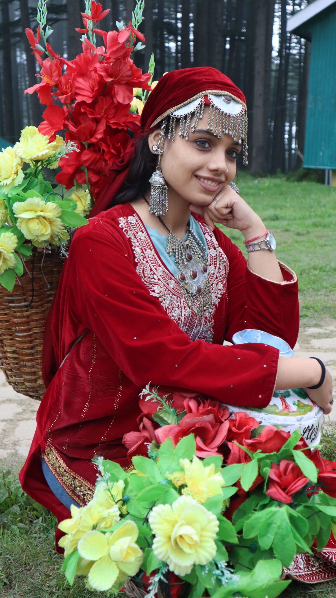 Young woman wearing traditional red velvet Himachali Chamba dress adorned with silver head jewelry, jhumkas, and neckwear near pine forests.