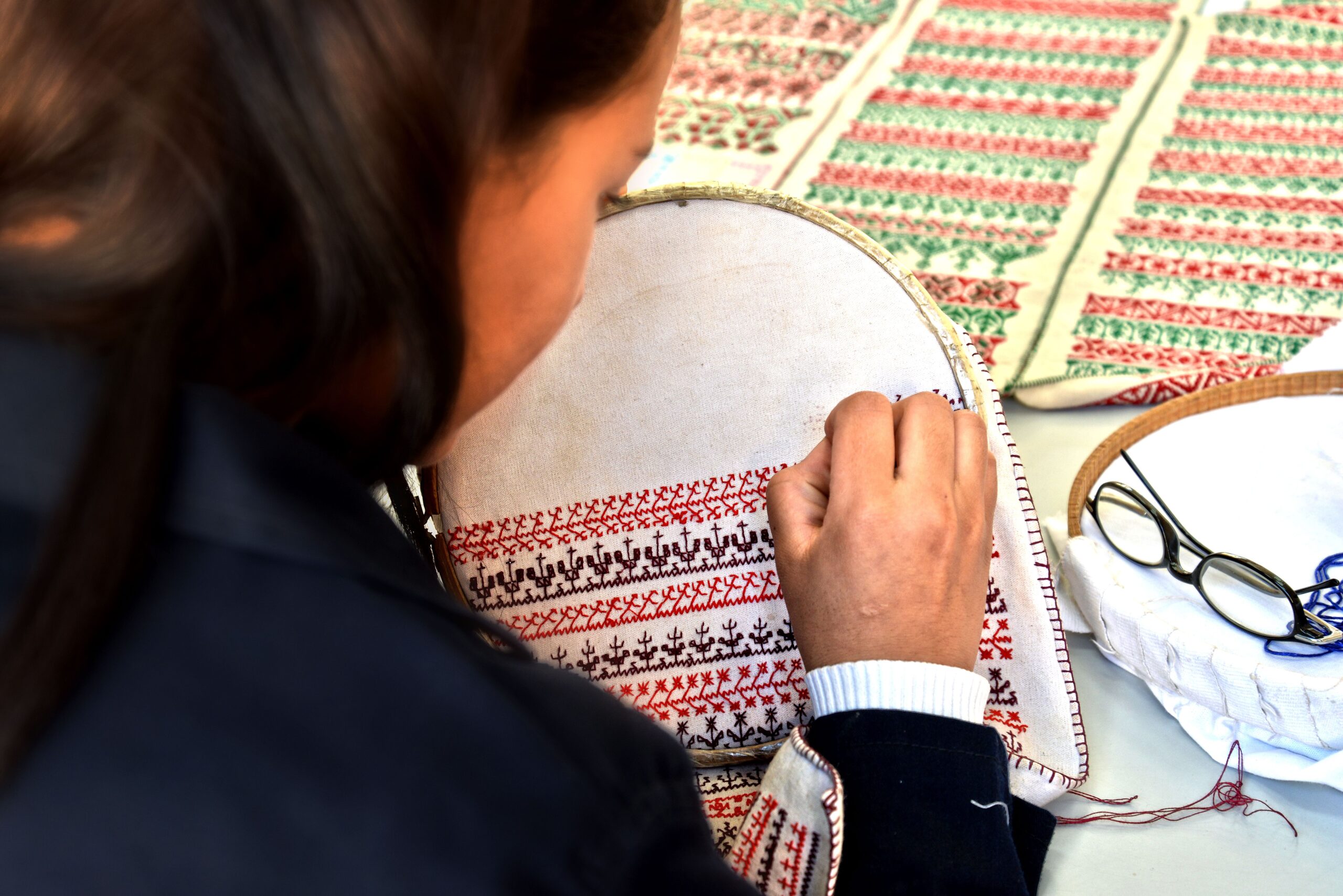 Artisan using a round wooden embroidery hoop to carefully hand-stitch traditional red and dark silk thread motifs on white fabric.